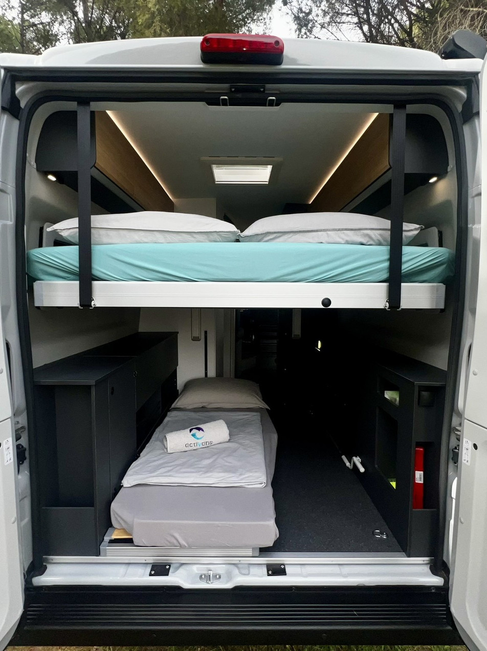 Camper mieten Spanien – Einzelbett Heckgarage mit Lattenrost | Adria Twin 640 SGX | Activans Blick von den geöffneten Hecktüren nach innen auf das Einzelbett am Boden unter dem Hauptbett mit Lattenrost im Adria Twin 640 SGX Camper