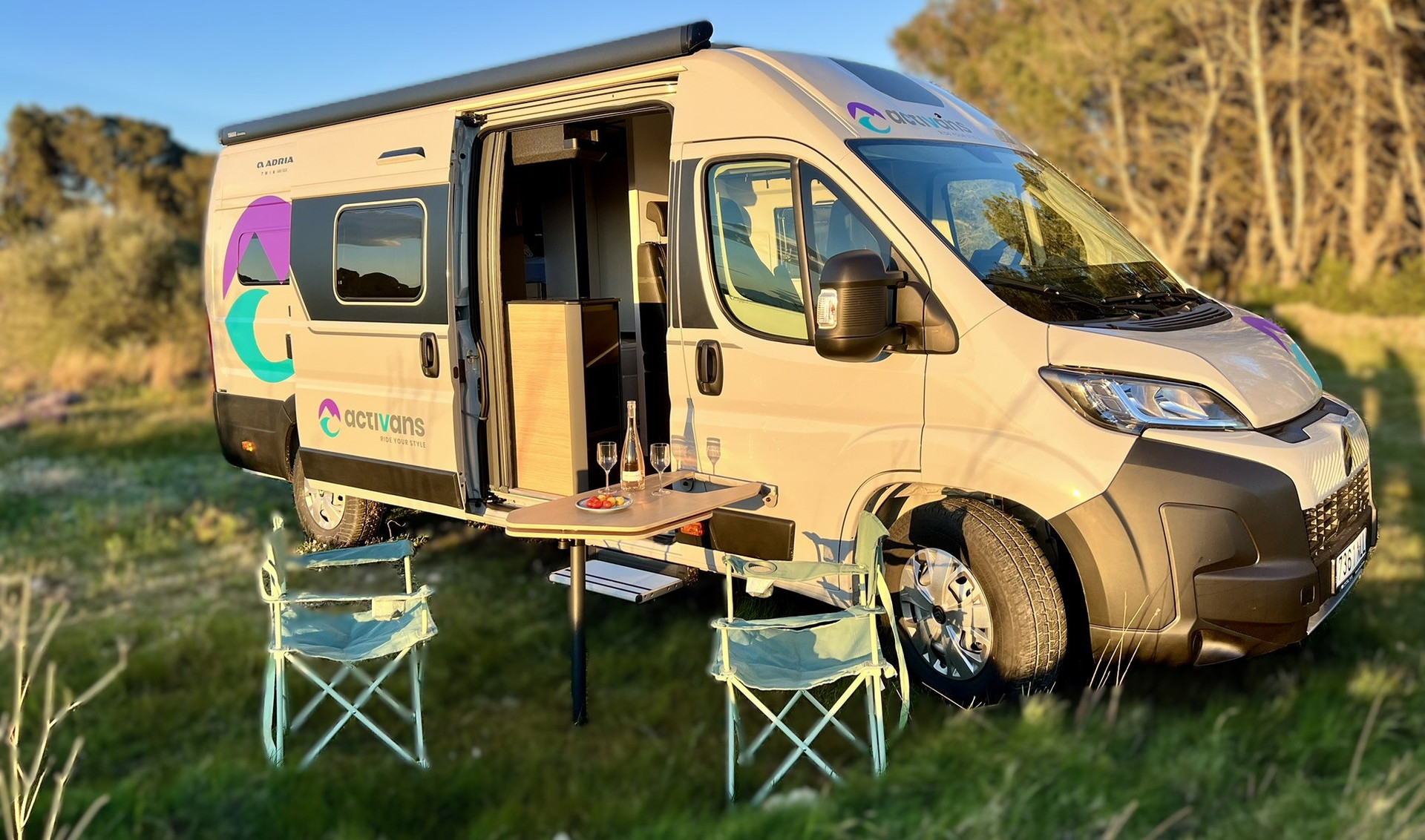 Alquilar camper Costa Brava – Adria Twin 640 SGX en la naturaleza al atardecer | Activans Camper Adria Twin 640 SGX con puerta lateral abierta, mesa, dos sillas, vino y aperitivo al atardecer en plena naturaleza – alquilar camper Costa Brava y Barcelona