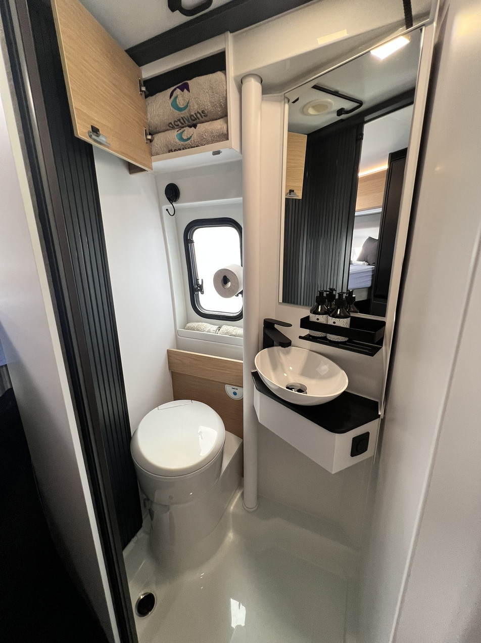 Baño pivotante amplio Adria Twin 640 SGX con armario abierto | Activans Baño pivotante amplio Adria Twin 640 SGX con toallas, lavabo y espejo grande – alquilar camper Barcelona