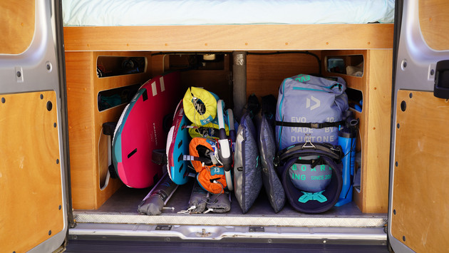 Vue à travers les portes arrière ouvertes sur la soute  du van qui est chargée d'équipement de planche à voile et de kitesurf.
