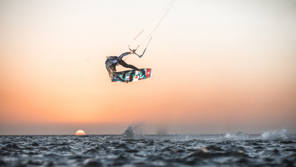 The sun rises in the background and a kitesurfer jumps in the air and touches the kiteboard with one hand