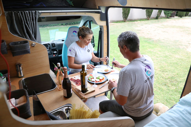 [Translate to Catalan:] 2 people sit at the table inside the camping van and eat.